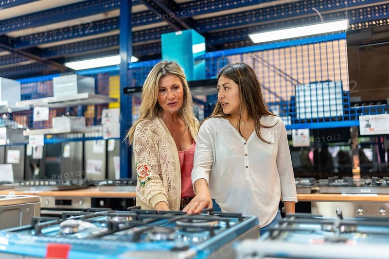 Mother and daughter compare gas cooktops in a retail appliance store, discussing kitchen renovation options and choosing equipment for their home upgrade and remodeling project