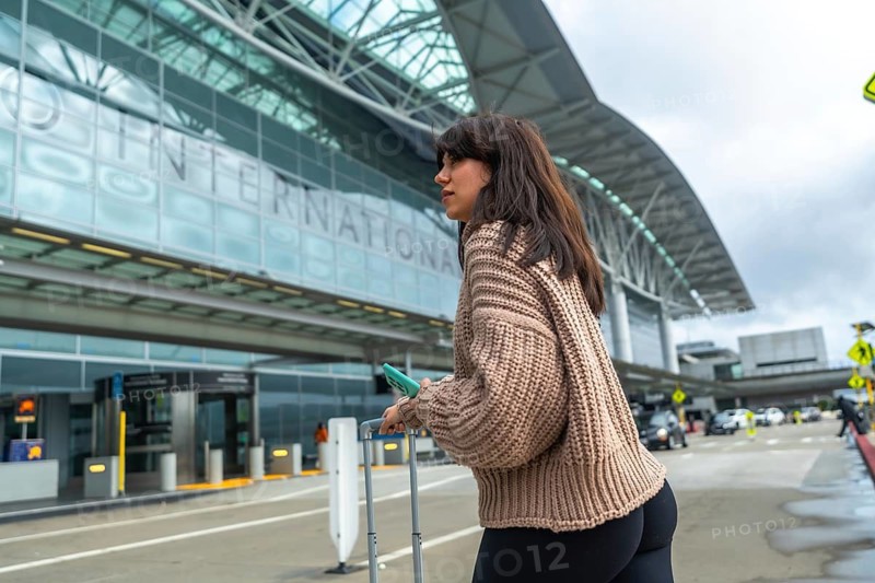 Young woman standing near the curb with luggage, holding a phone, and looking to the side, waiting for transportation or preparing for departure at an international airport building