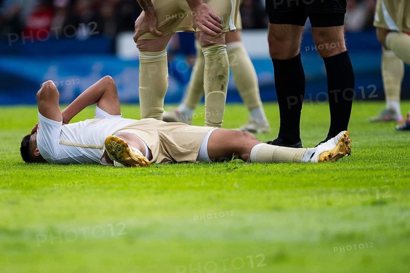 Injured football player lying on the pitch after a challenge while teammates and referee assess the situation during a match in Legnica, Poland