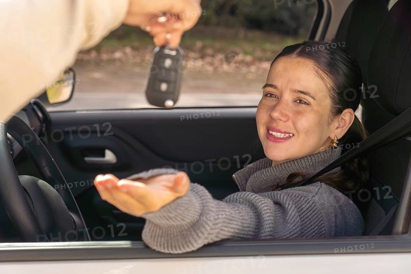 Smiling young woman looking at car key being handed to her through window, car interior, achievement concept