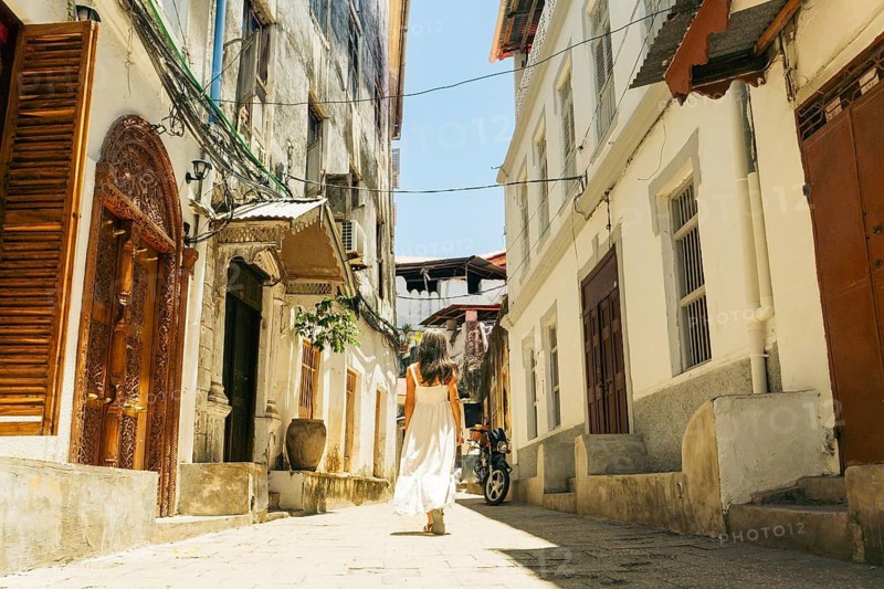 Woman tourist in white dress walks through beautiful Stone Town alley. Historic Zanzibar architecture and peaceful morning travel sightseeing.Popular travel destination Africa