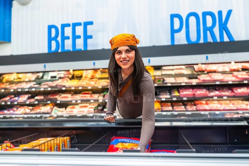Woman smiling, reaching for a package of frozen food in an open display freezer, with shelves of packaged meat products and beef and pork signage visible in the background