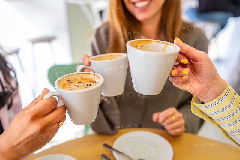 Three friends celebrating their friendship with a happy coffee toast, holding white cups filled with warm beverages and enjoying a social gathering at a bright cafe table