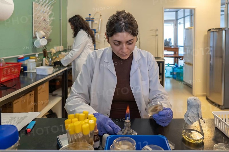 Young female scientist in white lab coat and blue gloves focused on working with a lit alcohol lamp and petri dish at a busy lab bench, with a colleague working in the background