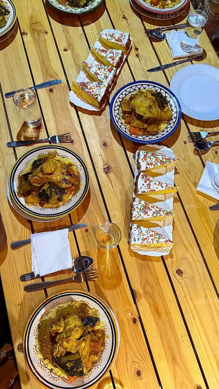 Delicious traditional moroccan meal with couscous, chicken tajine, assorted vegetables, and sweet cake slices with sprinkles arranged on a rustic wooden table, creating a festive dining scene