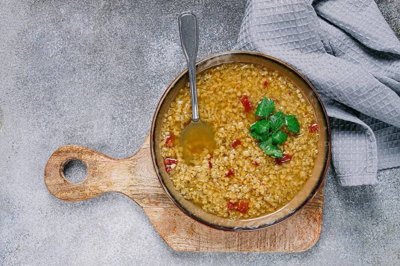 Savory lentil soup garnished with fresh herbs sits in a wooden bowl on a textured surface, accompanied by a silver spoon and a gray cloth napkin, bringing comfort and warmth