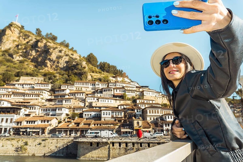 Smiling happy woman search gets perfect angle takes selfie shot, records vlog using her blue smartphone in Berat, Albania. Historic Ottoman houses of the Mangalem district. City of a Thousand Windows