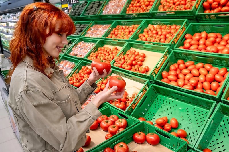 Young red haired woman smiling while selecting ripe red tomatoes from green crates in a bright supermarket produce section, choosing fresh, healthy food for cooking