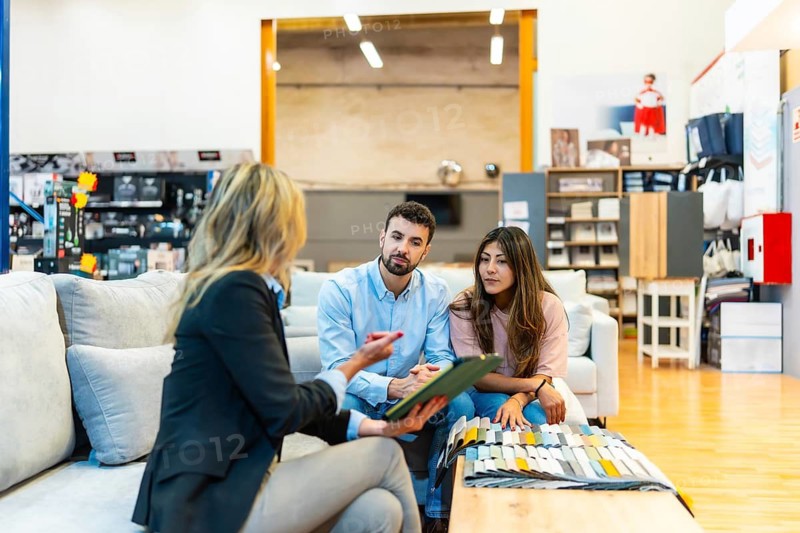 Saleswoman discussing material swatches and home decor options with a young adult couple while sitting in a furniture showroom, representing sales, client consultation, and design choices