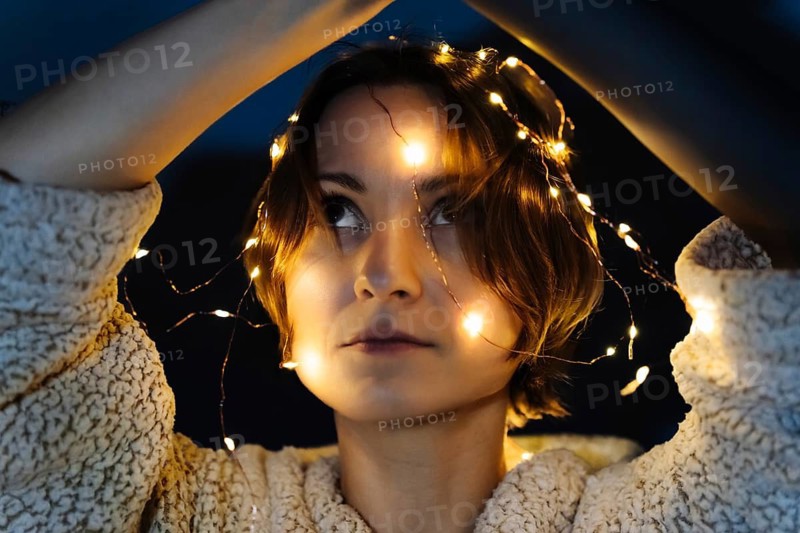 Portrait of a short-haired woman with string lights on the glove box. A woman wearing a light textured sweater raises her arms while warm fairy lights wrap around her head and shoulders against a dark background. The glowing lights and soft fabric create an intimate cozy mood suitable for winter lifestyle branding, holiday campaigns, emotional storytelling