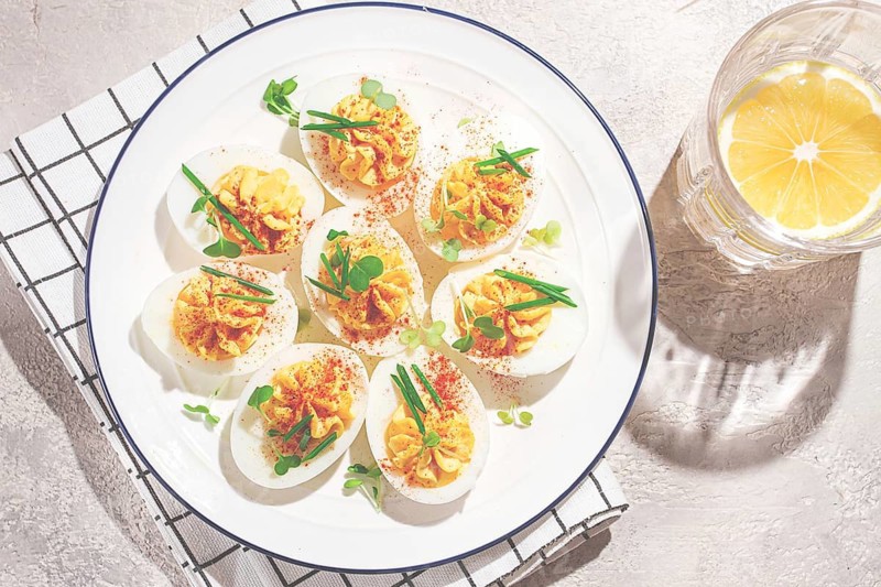 A plate holds several deviled eggs filled with yellow mixture and topped with spices and green garnishes. The dish is placed on a smooth light surface