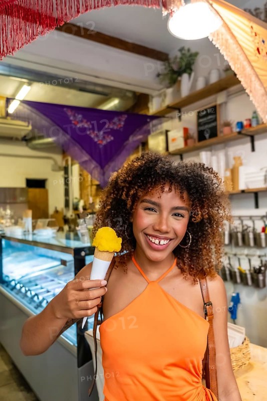 Young afro brazilian woman with curly hair smiling and holding a refreshing yellow ice cream cone, experiencing happiness and a sweet treat inside a brightly lit gelato shop