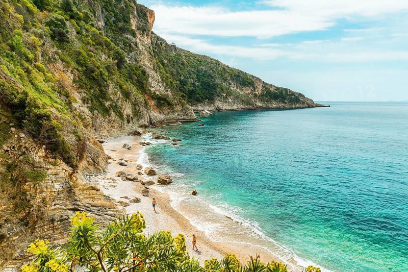 Viewpoint panorama couple enjoy walk isolated on Filikuri beach in spring. Clear turquoise Ionian sea meets steep rocky coastal cliffs. Scenic Albanian Riviera travel landscape in Himar, Albania