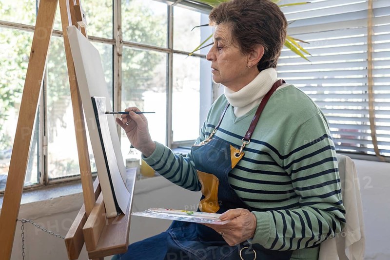 Older female artist applies brushstrokes to a canvas while holding a watercolor palette and working at a wooden easel in a bright home studio filled with natural light
