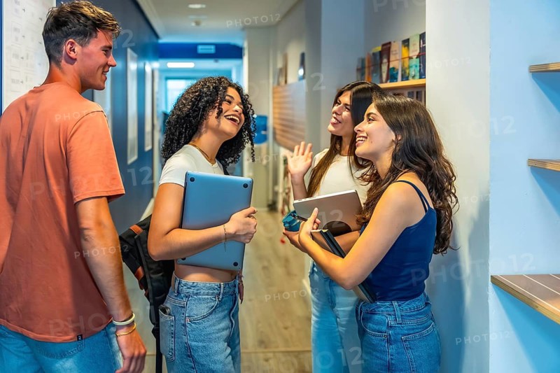 Group of happy young students standing outside a classroom, holding laptops and tablets, casually chatting and smiling during a break in a university or college hallway