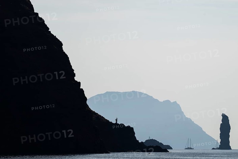 Dramatic cliffs and silhouette of a towering rock pin, behind it the island of Alicudi, a sailboat in the calm sea, near Filicudi, Aeolian or Lipari Islands, southern Italy, Italy