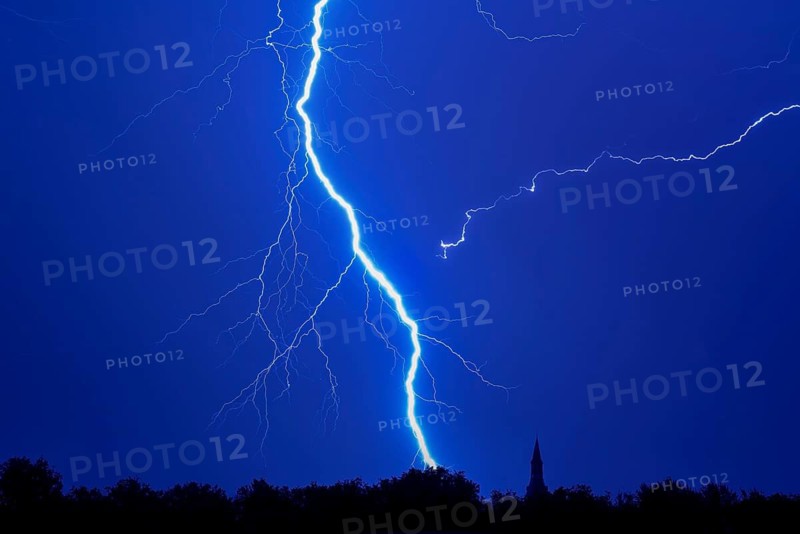 Forked lightning bolt, cloud-to-ground lightning strike during thunderstorm in blue night sky over forest with silhouetted treetops and church tower