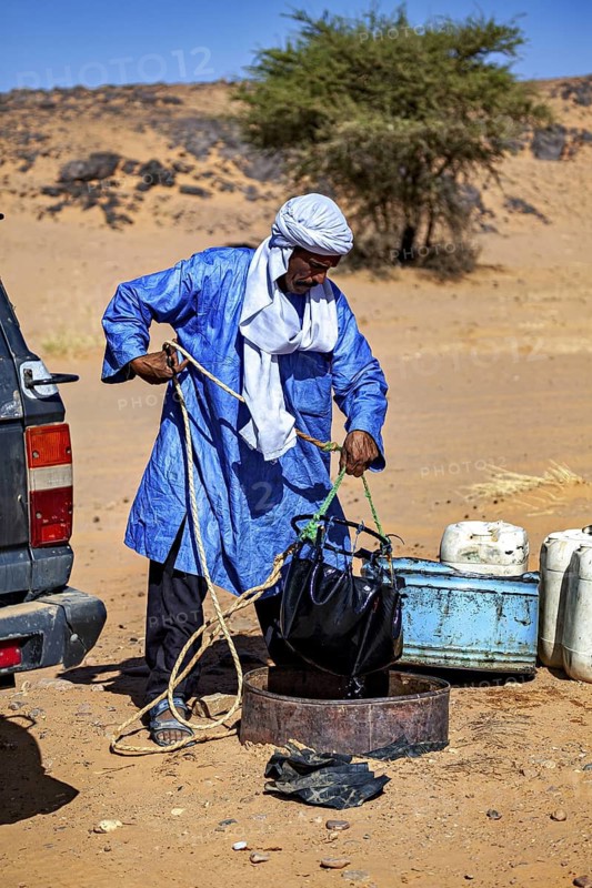 A man in a blue robe deals with a water container in a desert-like environment, Tuareg draw water from a well to wash themselves in the Sahara in Algeria