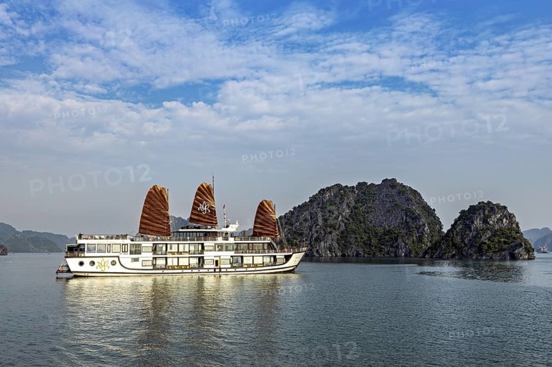 Boat with sails on calm sea in front of limestone islands, blue sky and clouds, cruise ship for tourists in Halong Bay in Vietnam