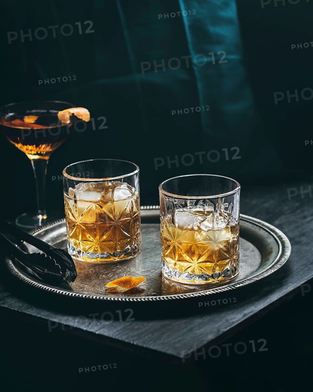 Two whiskey glasses filled with ice rest on a silver tray alongside a cocktail featuring a vibrant orange garnish, set against a dark and elegant background