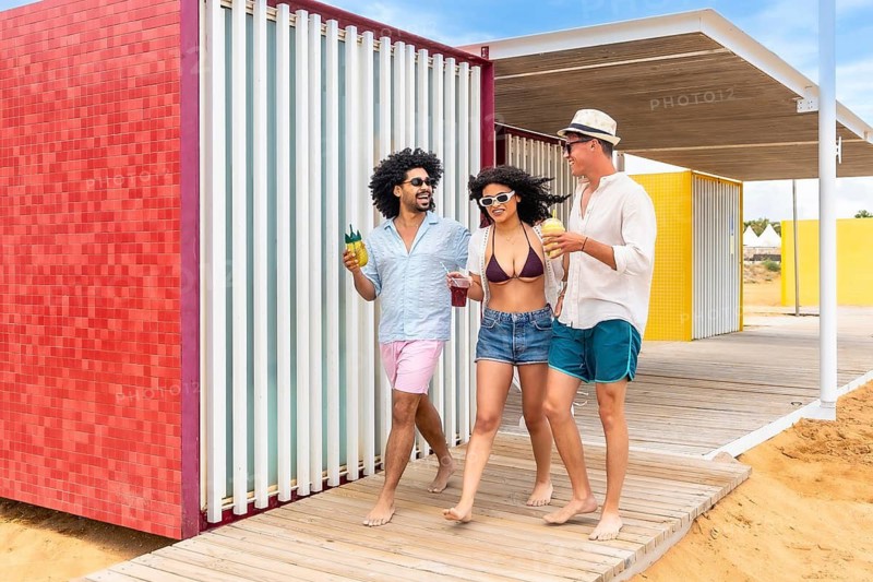 Diverse friends walking barefoot on a sunny beach boardwalk, laughing and sipping cold drinks beside colorful modern beach architecture, enjoying a carefree summer getaway