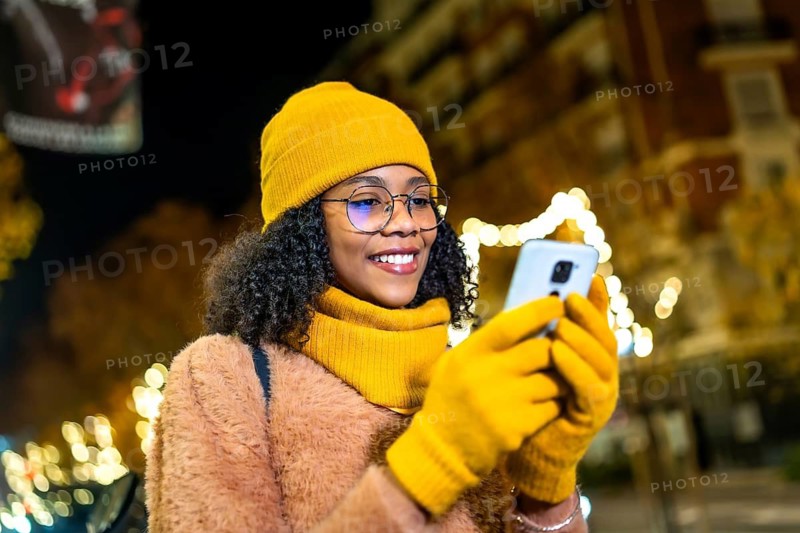 Smiling young woman wearing yellow winter clothes and gloves using smartphone in a city street at night with christmas lights