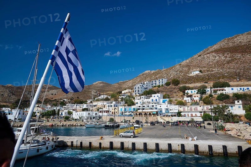 Greek flag flying over a harbor, with white coastal town in the background, Zero Waste Island, Livadia, Tilos, Dodecanese, Greek Islands, Greece