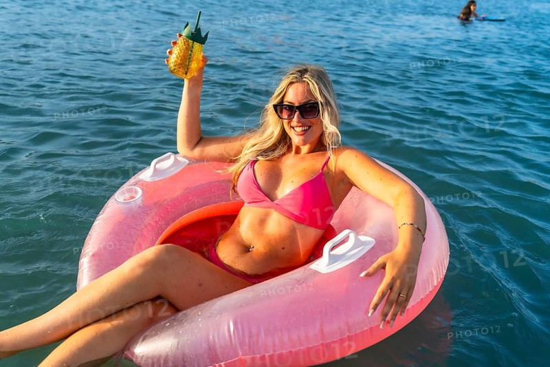 Young woman smiling while floating in the ocean on a pink inflatable ring, holding a tropical drink and enjoying her summer vacation