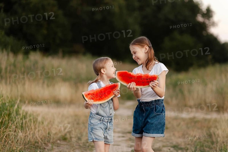Two children smiling and holding watermelon slices outdoors on a summer day. They stand in a grassy field, embracing the joys of summer and friendship together