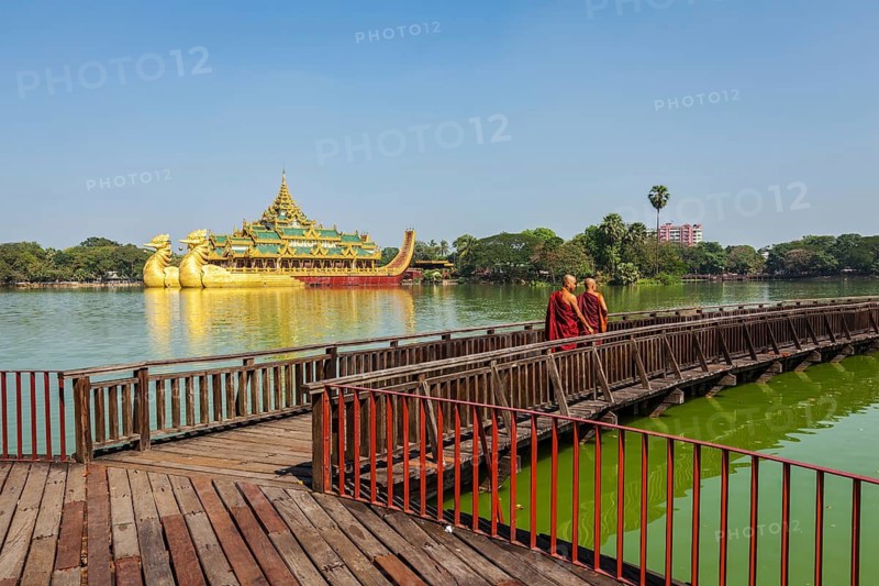 YANGON, MYANMAR, JANUARY 3, 2014: Two Buddhist monks walking on wooden bridge at at Kandawgyi Lake with Karaweik barge in Background