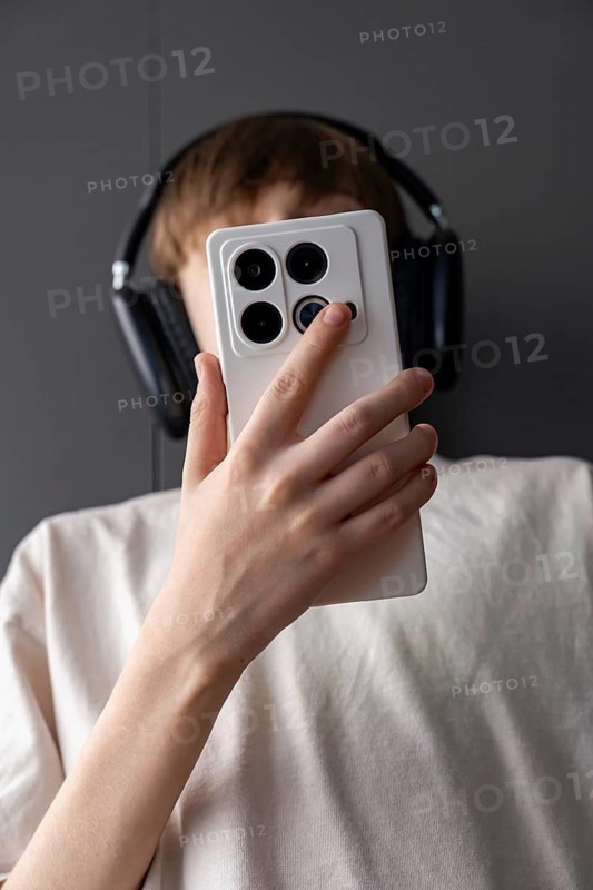 A teen wearing large black headphones holds a smartphone with a white case up in front of their face. The close-up captures a modern moment of digital interaction, focus, or content creation