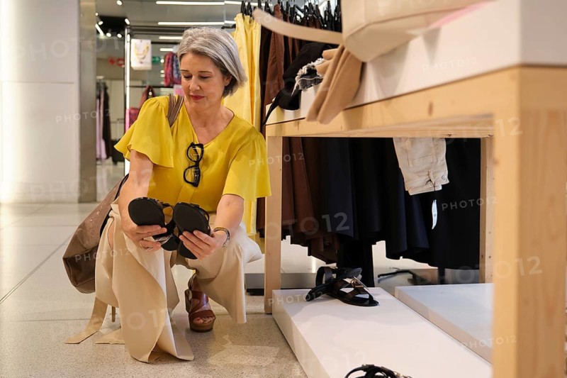 A mature woman is seen in a stylish store, kneeling and examining a pair of fashionable black sandals. She appears thoughtful, comparing options for purchase