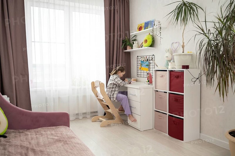A young girl sits barefoot at her desk in a bright, organized bedroom, drawing on a digital tablet. Surrounded by soft decor, storage, and natural light, she enjoys a creative and focused moment