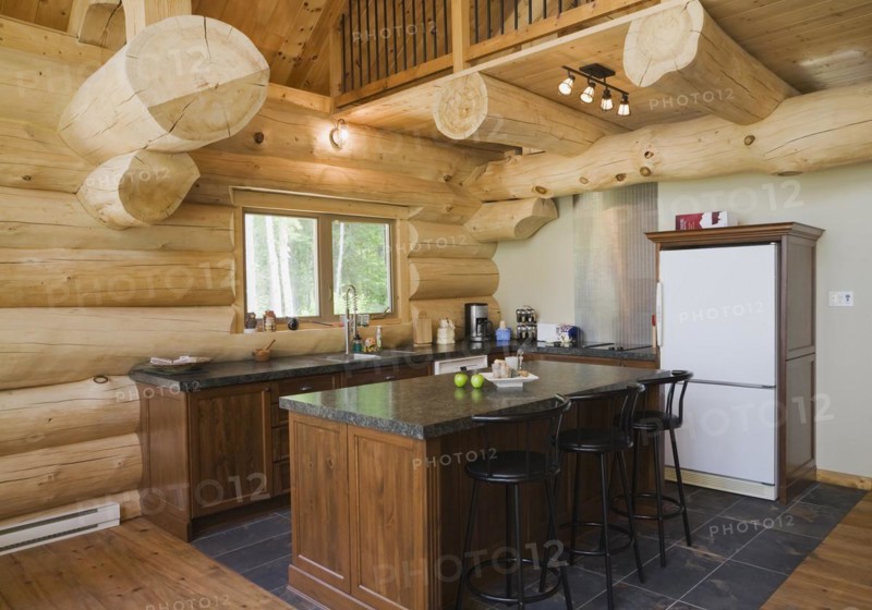 Island with dark grey granite countertop and black stained wooden bar stools in kitchen with ceramic tile floor inside handcrafted Eastern white pine Scandinavian log cabin home, Quebec, Canada