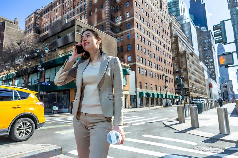 Businesswoman walking on a city street, communicating on her cell phone and holding a reusable coffee cup, representing urban life, connectivity, and a busy corporate commute in new york city