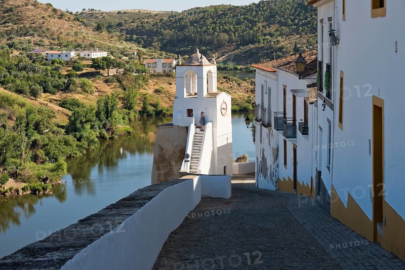 A woman in a blue summer dress stands on the clock tower (Torre do Relogio), which was built in one of the towers of the city walls on the right bank of the River Guadiana. Mertola. Baixo Alentejo. Portugal
