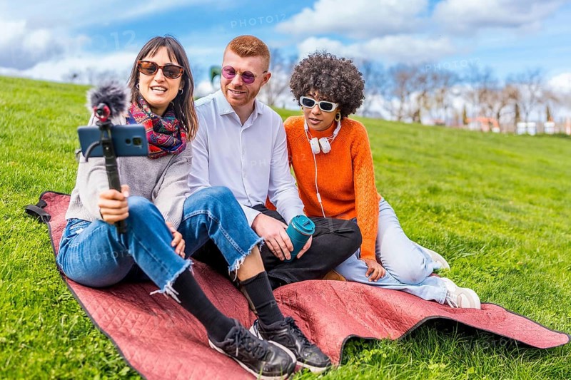 Three content creators filming a video blog in a park using a smartphone with external microphone, sitting on a blanket