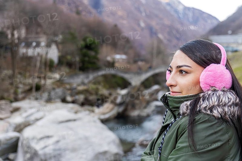 A woman with pink earmuffs gazes at the picturesque Ponte Dei Salti bridge in Lavertezzo, Ticino, Switzerland. The winter landscape with rocky riverbed creates a serene backdrop