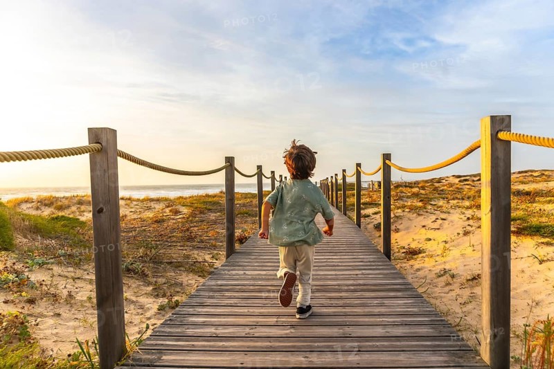 Little boy running fast along a wooden boardwalk towards a sandy beach and ocean, enjoying a summer vacation at sunset with clear skies in seignosse, france