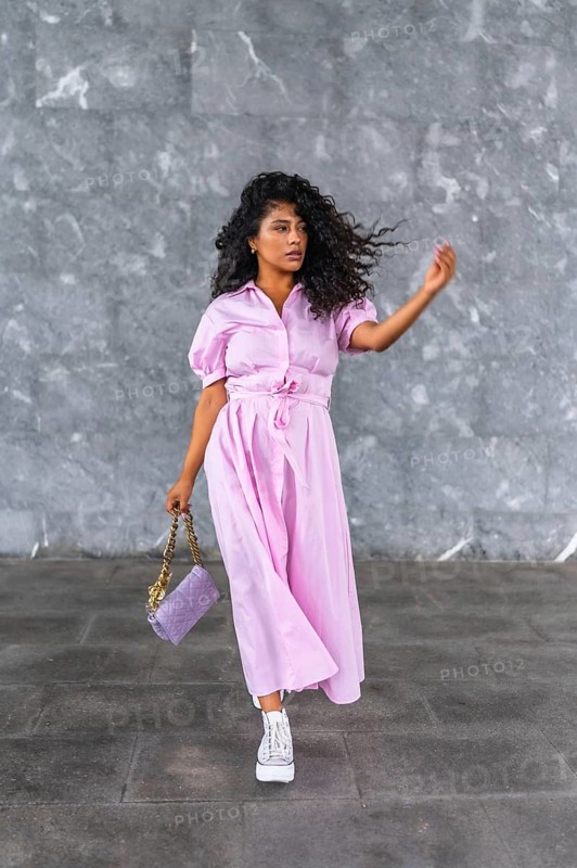 Vertical close-up portrait against a grey urban wall of a latin beauty model walking wearing pink clothes and holding purse
