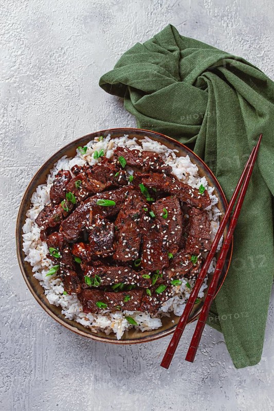 Crispy beef, with green onions and sesame seeds, on a rice pad, in a bowl with chopsticks, on a textured surface, no people