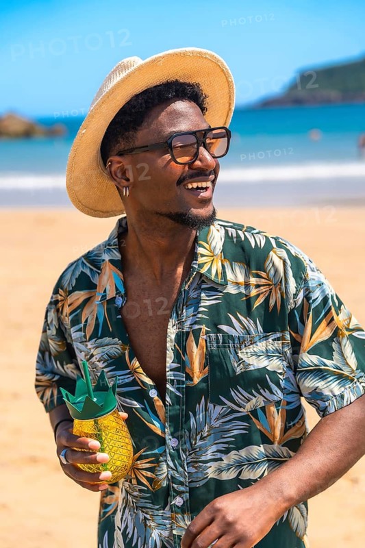 Young man wearing stylish sunglasses and a trendy hat, savoring a refreshing cocktail while relaxing on a sunny beach