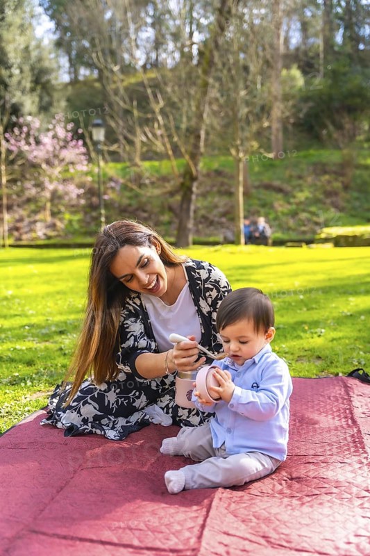 Happy mother feeding her baby boy solid food from a thermos while having a picnic in a park