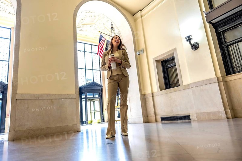 Young professional woman entering an arched hallway, holding a take out coffee cup, smiling and looking up, with an american flag visible in the background
