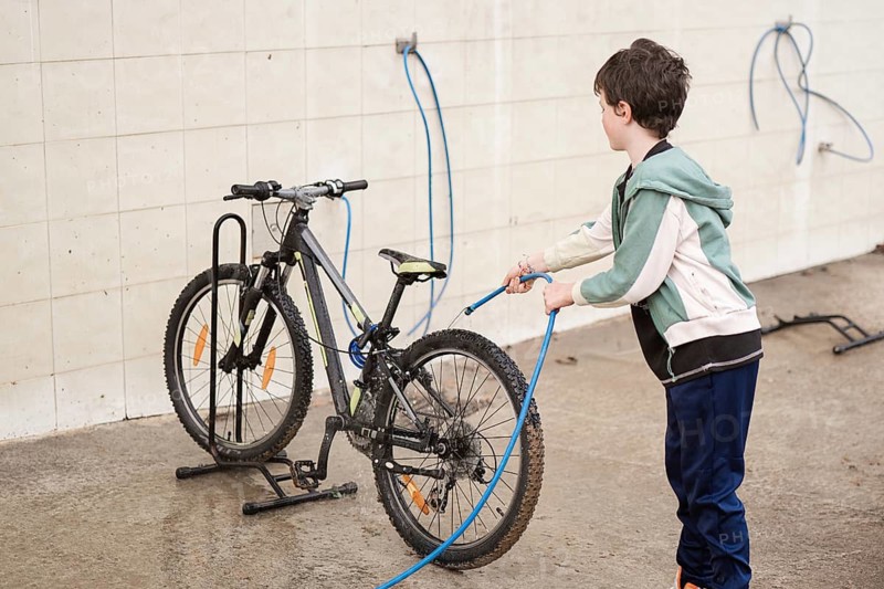 A young child attentively cleaning a bicycle with a hose in an outdoor space. The setting emphasizes responsibility, independence, and the enjoyment of outdoor activities