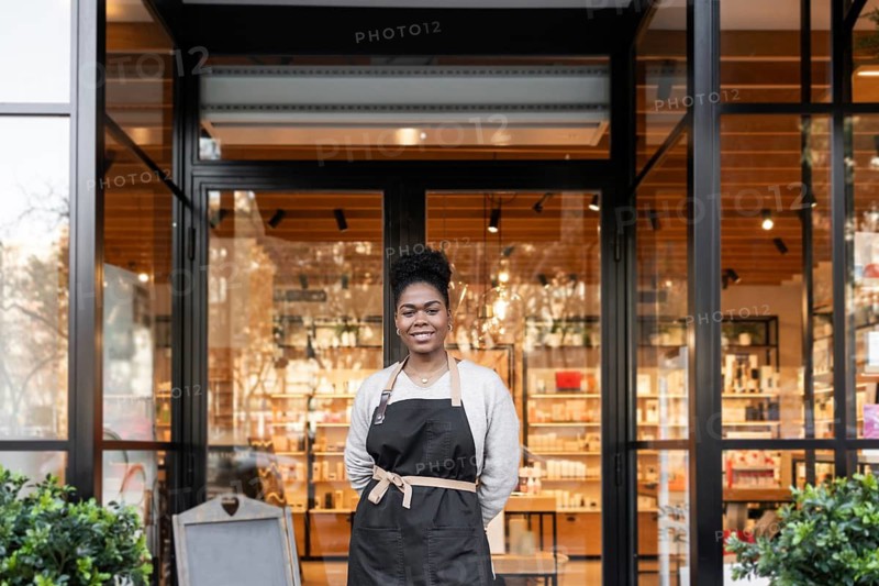 A friendly worker stands confidently in front of a vibrant cosmetics store, wearing an apron. The store's modern interior is visible through large glass windows, displaying a variety of products
