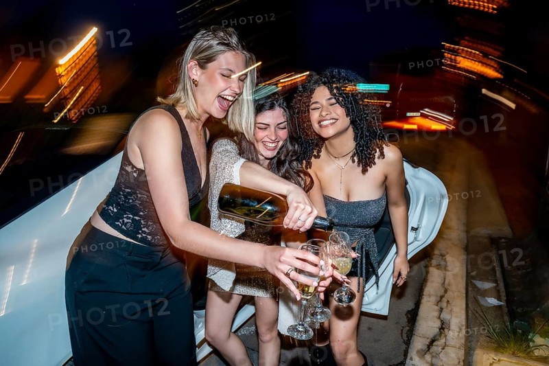 Three cheerful young women in a limo celebrate a glamorous night out, smiling and pouring sparkling wine as they toast, laugh, and enjoy friendship and luxury
