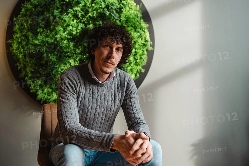 Curly-haired young man sitting in front of a lush green wall. The setting emphasizes themes of ecology, sustainable development, and environmental responsibility