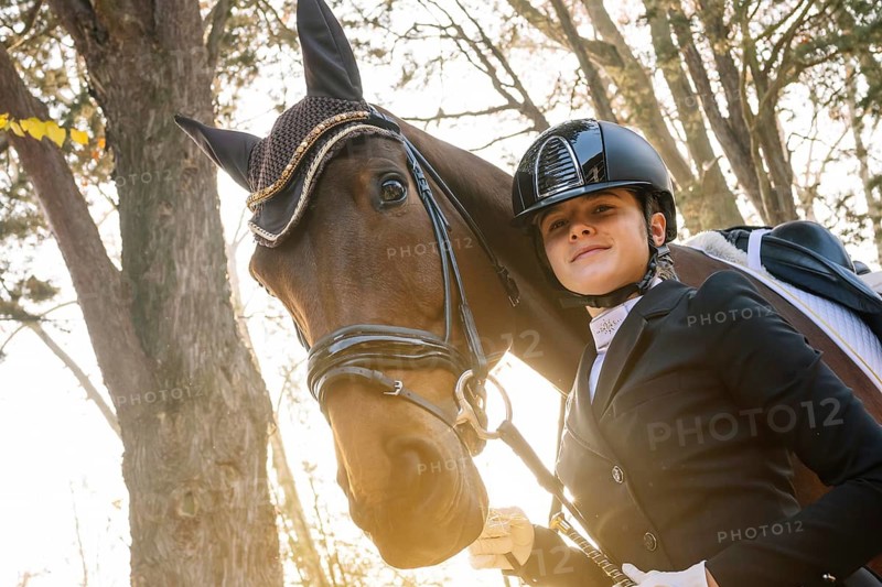 Young equestrian in classical dressage attire stands confidently beside her horse Sunlight filters through autumn trees, highlighting the bond between rider and horse