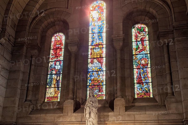 Three colourful windows with a statue in front of them, sunlight streaming through the middle window, Paris